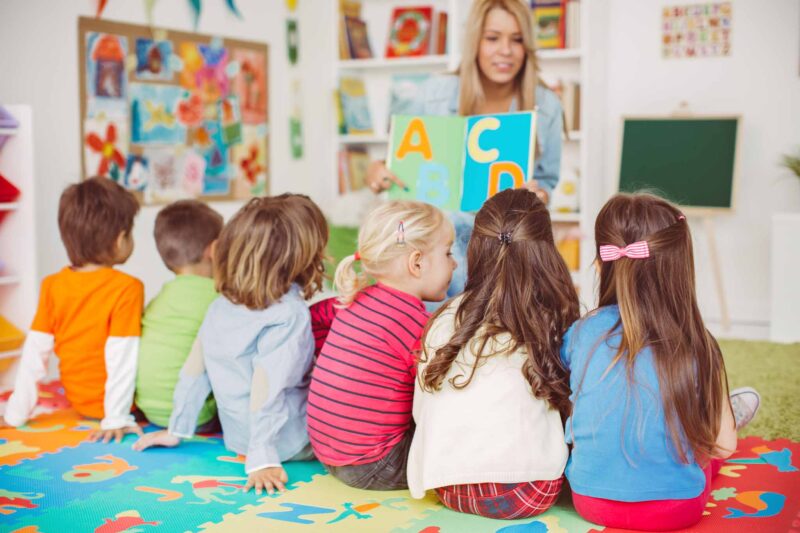 Teacher with a group of preschool children in a nursery. The children are sitting on the floor and listening teacher. Learning letters. In the background we can see a shelf with some, toys, black board and books. View from behind.