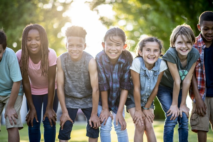 A group of kids are outdoors on a sunny day. They are posing with their hands on their knees for a fun group photo.