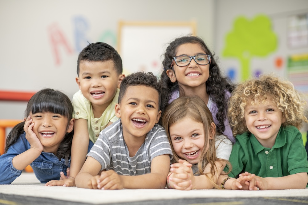 A small group of 6 Kindergarten students lay stacked on top of one another, on their classroom floor, as they pose for a portrait. They are each dressed casually and are smiling for the photo.
